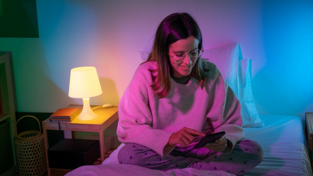 Schöne junge Frau sitzt im Bett und schaut auf ihr Handy, spielt mit den intelligenten Lichtern im Hintergrund