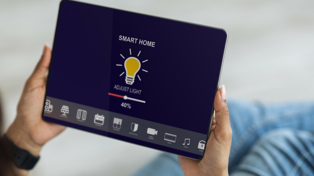 Eine Frau hält ein Tablet in den Händen, auf dem eine Smart-Home-App geöffnet ist. Sie stellt die Helligkeit der intelligenten Glühbirnen in ihrer Wohnung ein